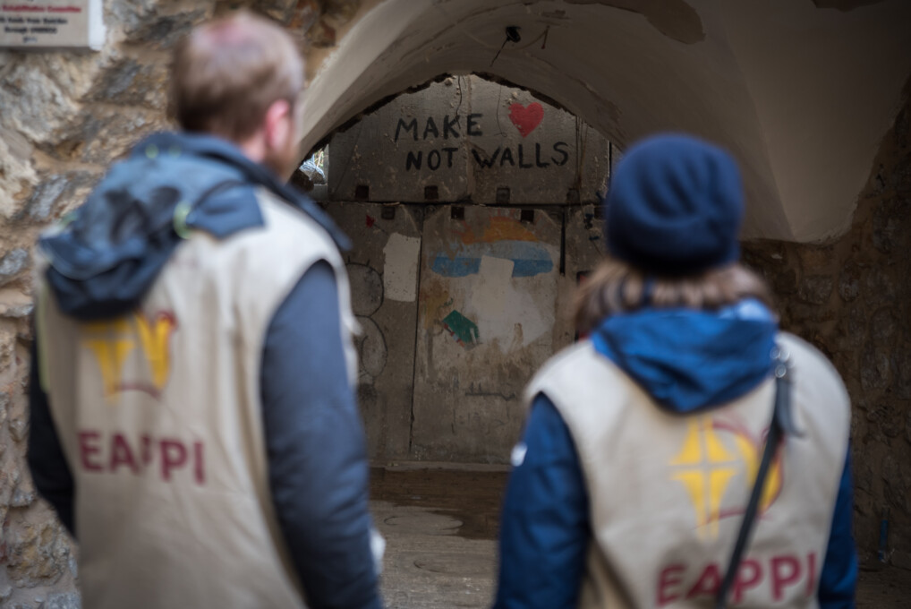 To EAPPI-ledsagere står med ryggen til kameraet. De kigger på en mur, på hvilken der med graffiti står skrevet: "Make Love, Not Walls". Muren separerer souq'en i Hebrons gamle bydel fra en israelsk bosættelse inde i byen.
