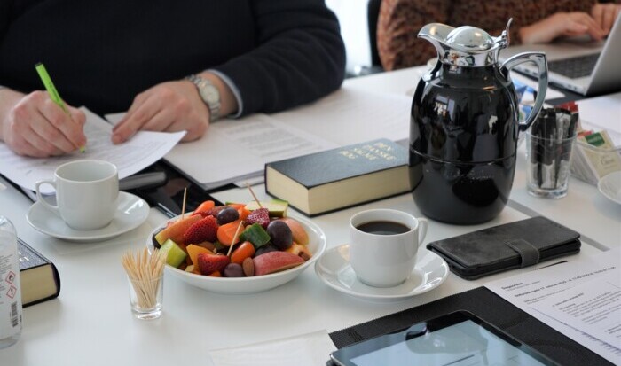 Genrebillede af stiftsrådsmøde, menighedsrådsmøde eller lignende. På billedet ses hænder, frugt, kaffekop, salmebog, etc.